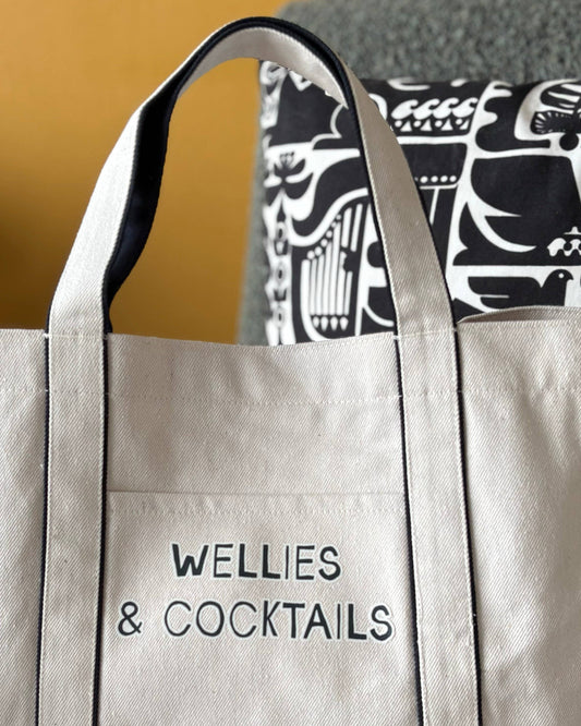 Beige tote bag with 'Wellies & Cocktails' text, worn over a patterned garment.
