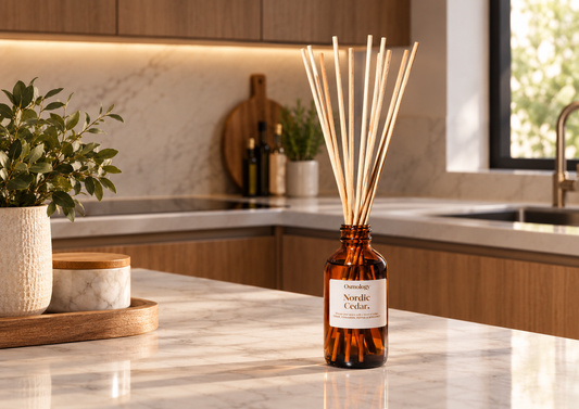 An amber glass reed diffuser bottle with a white label reading 'Gentle Nordic Cedar' sits on a marble kitchen counter. The diffuser has light natural reeds and is styled in a warm, modern kitchen setting with wooden cabinets, a potted plant, and a window.