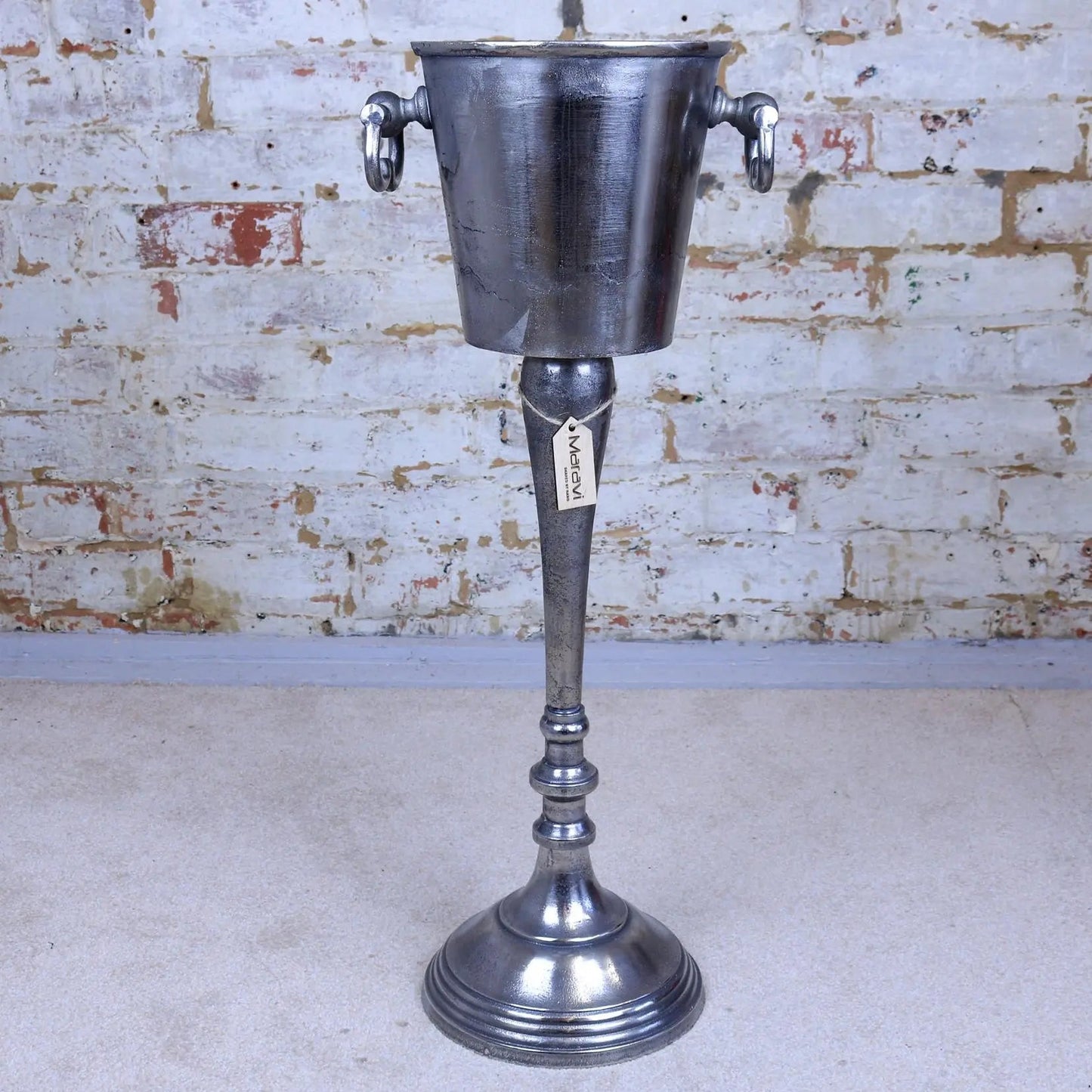 Vintage-style metal ice bucket on a stand against a brick wall.