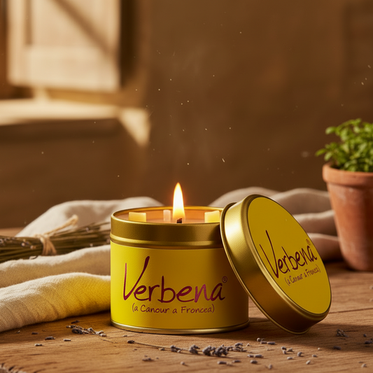 A yellow and gold metal candle tin labeled 'Verbena (la Cour a France)' with its lid leaning against the open tin, set on a wooden surface with a lit candle flame, draped fabric, dried herbs, and a small potted plant in the background.