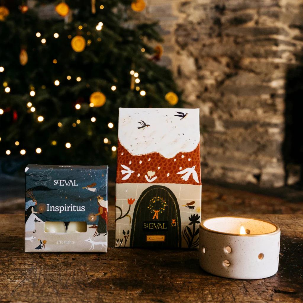 Two candles in decorative boxes with a lit candle on a wooden surface, Christmas tree and fireplace in the background.
