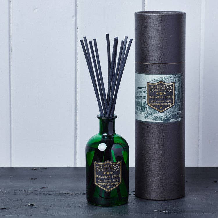 A reed diffuser bottle made of dark green glass with black reeds, standing next to its tall dark gray cylindrical packaging tube. Both pieces feature a gold and white label for 'The Regency Collection' and 'Palafrina Spice'.