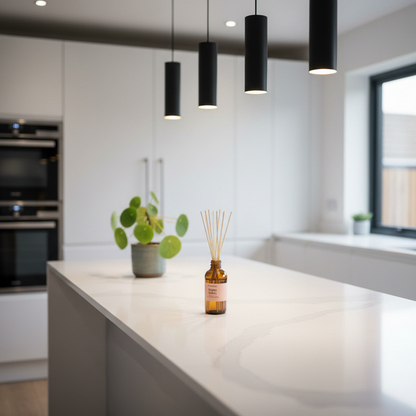 A reed diffuser bottle with wooden reeds sits on a white kitchen island, with a potted plant and black pendant lights in the softly blurred background.