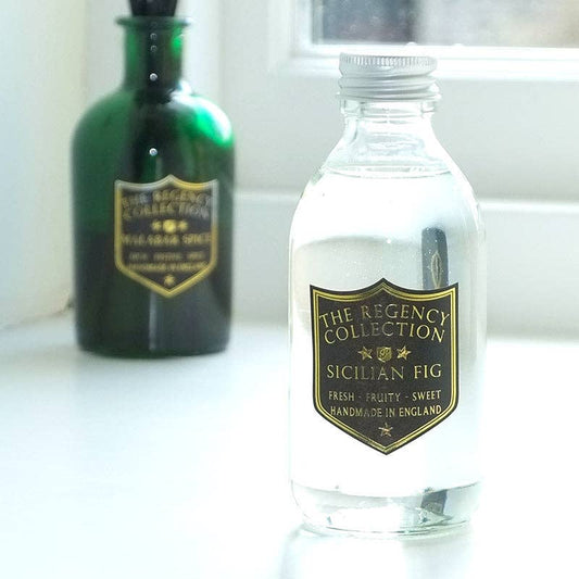 A clear glass bottle labeled 'The Regency Collection Sicilian Fig' in the foreground on a white windowsill, with a dark green bottle blurred in the background.