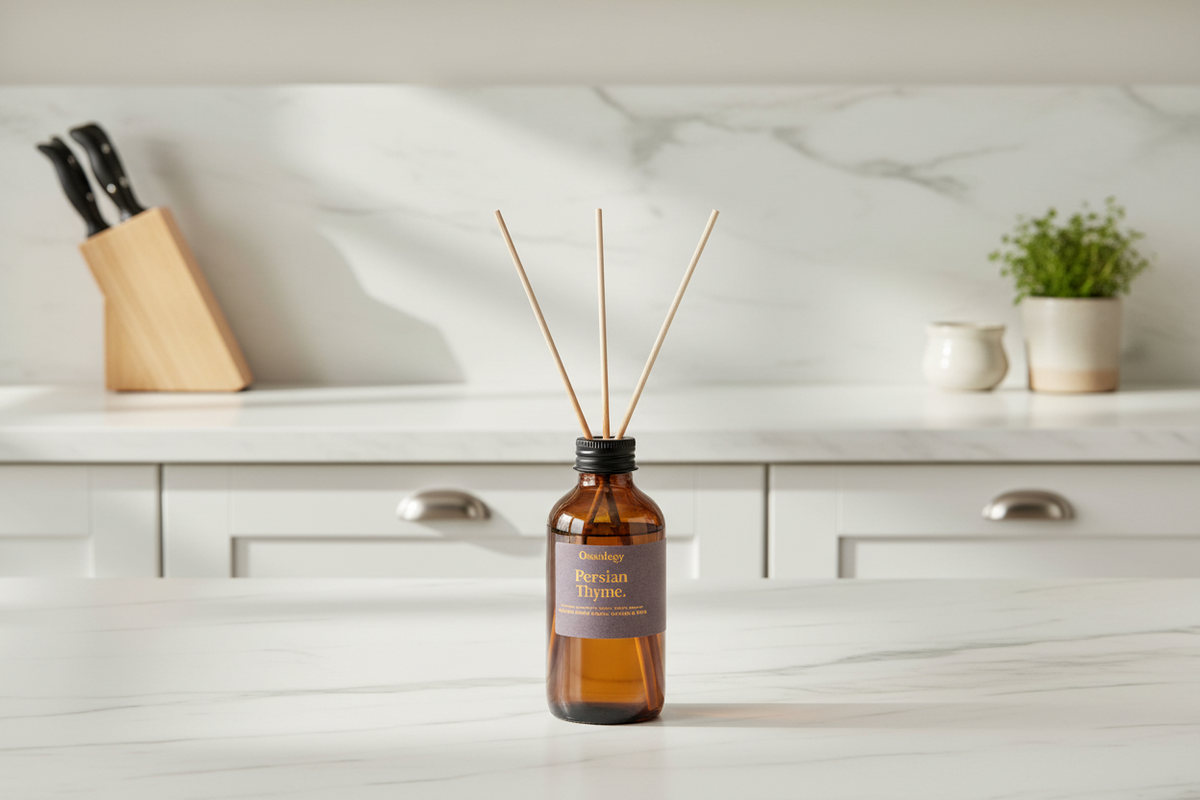 Amber glass reed diffuser bottle with three wooden reeds and a dark label reading 'Persian Thyme', placed on a white marble countertop in a softly blurred kitchen setting.