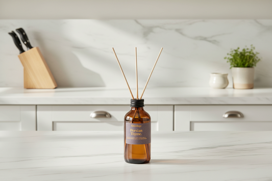 Amber glass reed diffuser bottle with three wooden reeds and a dark label reading 'Persian Thyme', placed on a white marble countertop in a softly blurred kitchen setting.