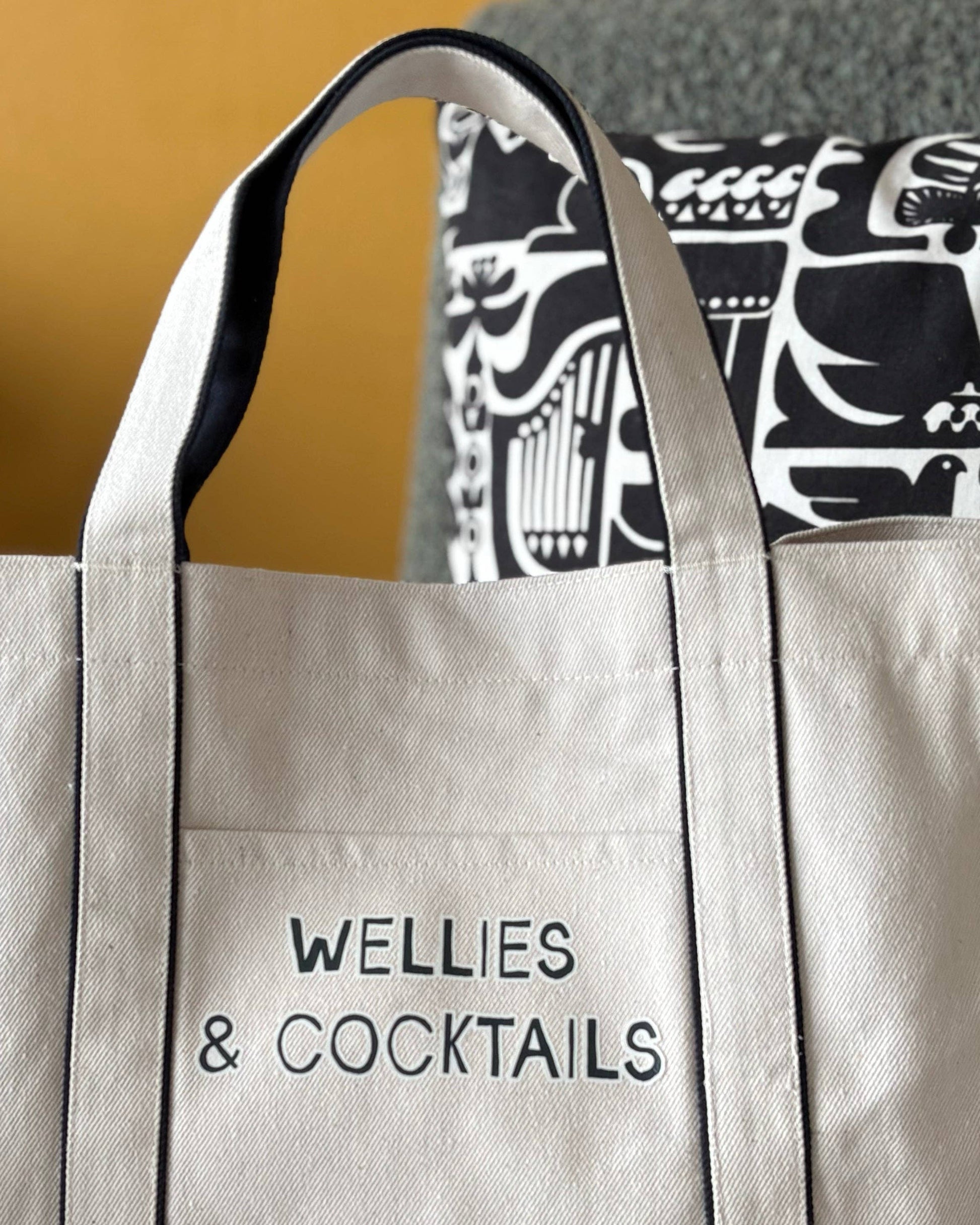 Beige tote bag with 'Wellies & Cocktails' text, worn over a patterned garment.