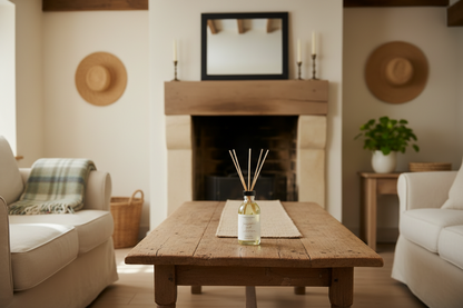 A rustic wooden coffee table in a cozy living room setting, holding a clear glass reed diffuser bottle with reeds. The room features a stone fireplace, cream-colored sofas, and straw hats on the walls.