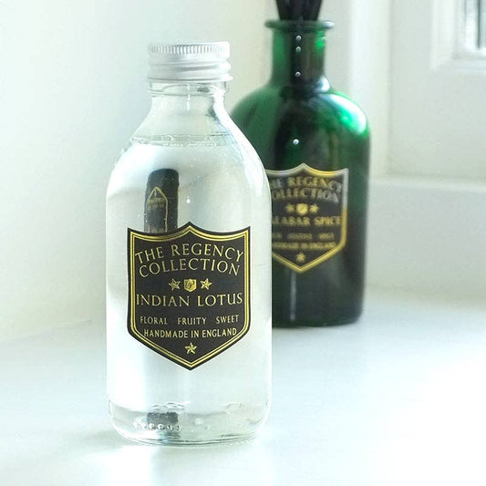 A clear glass bottle of The Regency Collection Indian Lotus liqueur in the foreground, with a dark green glass bottle of the same brand in the background, both on a white surface.