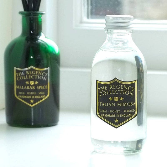 Two decorative bottles on a white surface near a window: a dark green glass reed diffuser bottle labeled 'The Regency Collection Malabar Spice' and a clear glass bottle labeled 'The Regency Collection Italian Mimosa' with white liquid and a silver cap.