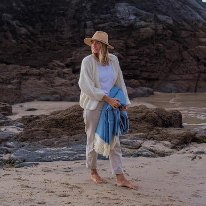 Woman standing on a rocky beach holding a blue towel, wearing a beige hat and light clothing.