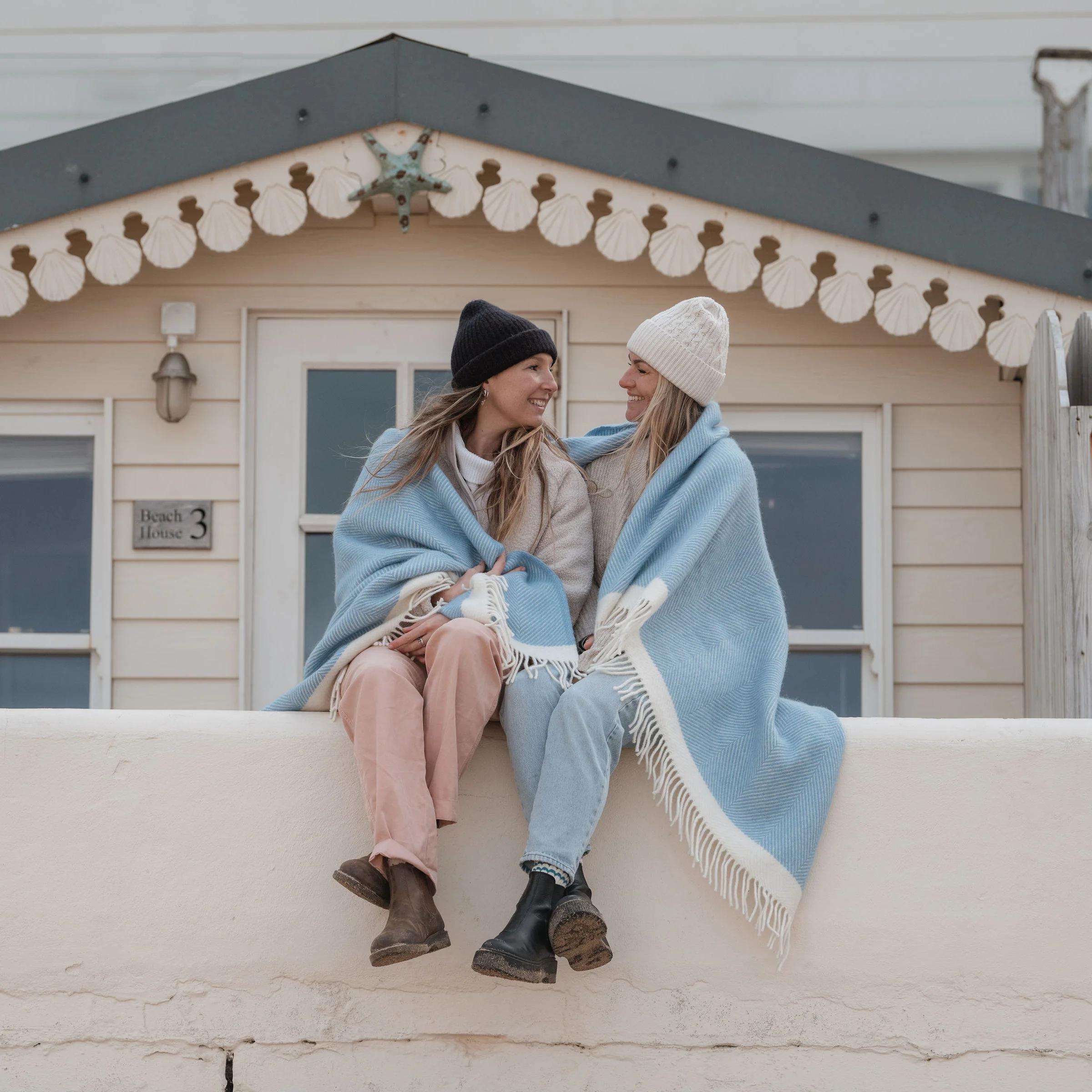 Two people sitting on a wall wrapped in a blue blanket with a beach hut in the background.