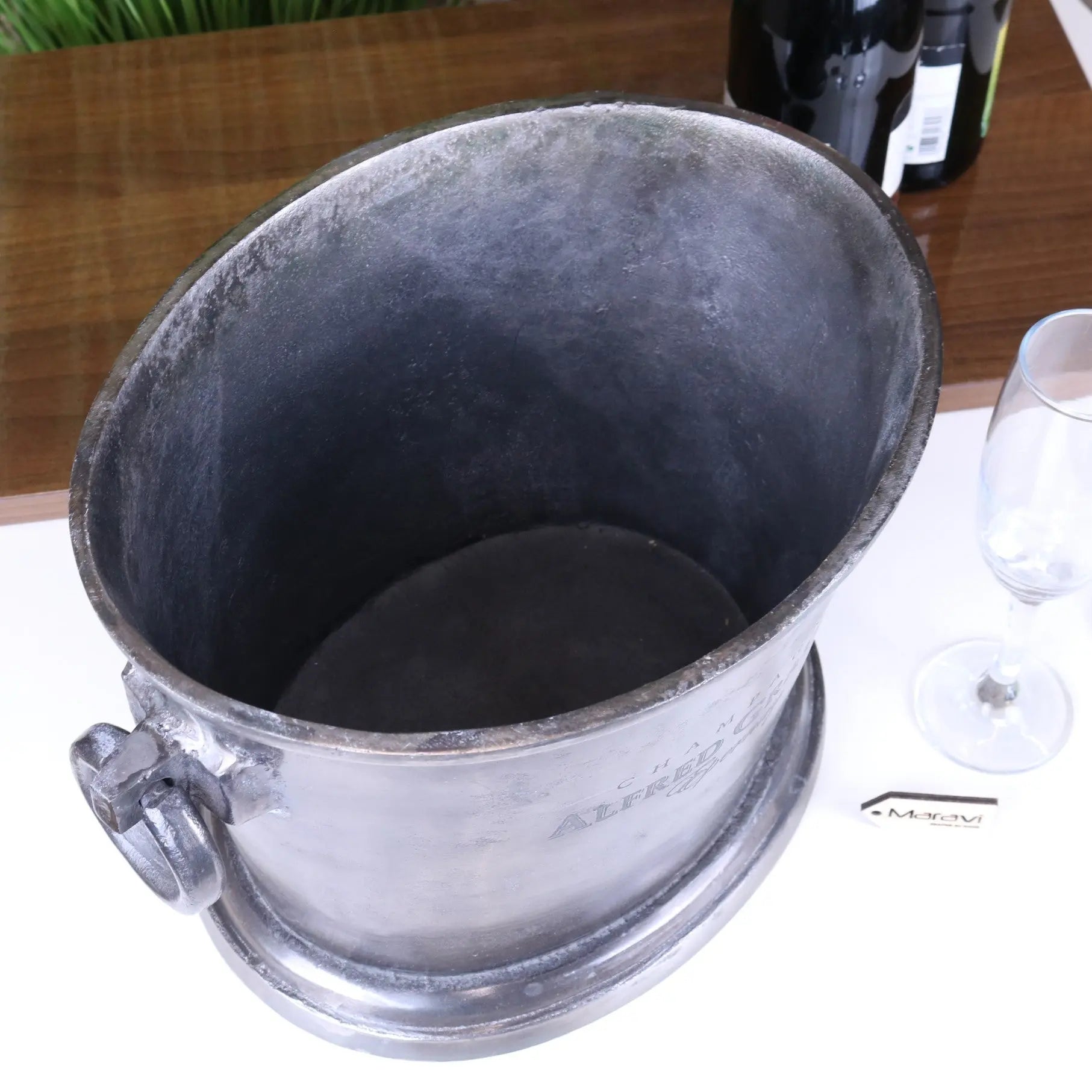 Metal wine cooler bucket on a table with a glass of water and a bottle in the background.