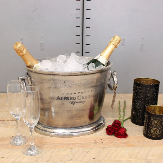 Silver champagne cooler with two bottles of champagne, glasses, and roses on a wooden table.