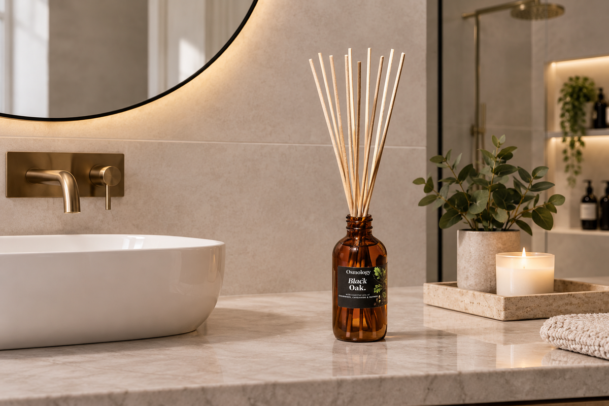 Amber glass reed diffuser bottle with wooden reeds on a bathroom countertop beside a white vessel sink, with a lit candle and small plant in the background.