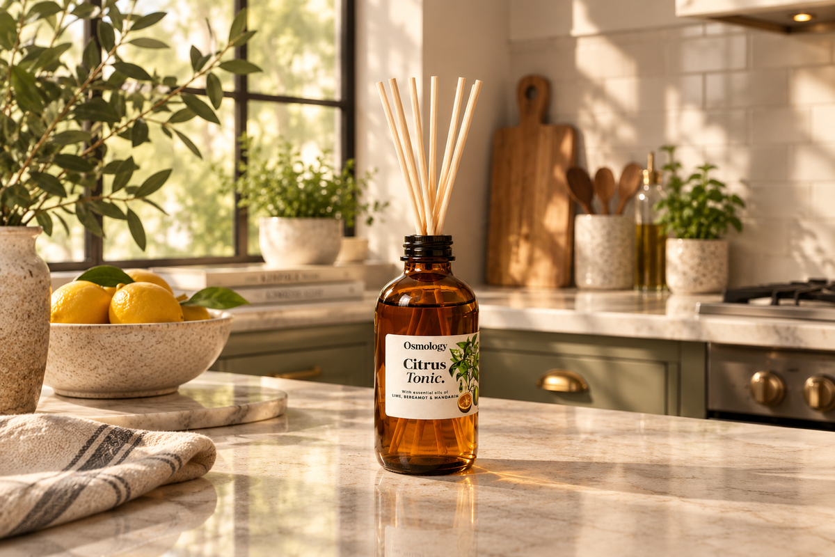 An amber glass reed diffuser bottle labeled 'Osmotik Citrus Tonic' stands on a marble kitchen counter with wooden reeds inserted, surrounded by lemons, plants, and kitchen decor in a bright, sunlit kitchen.