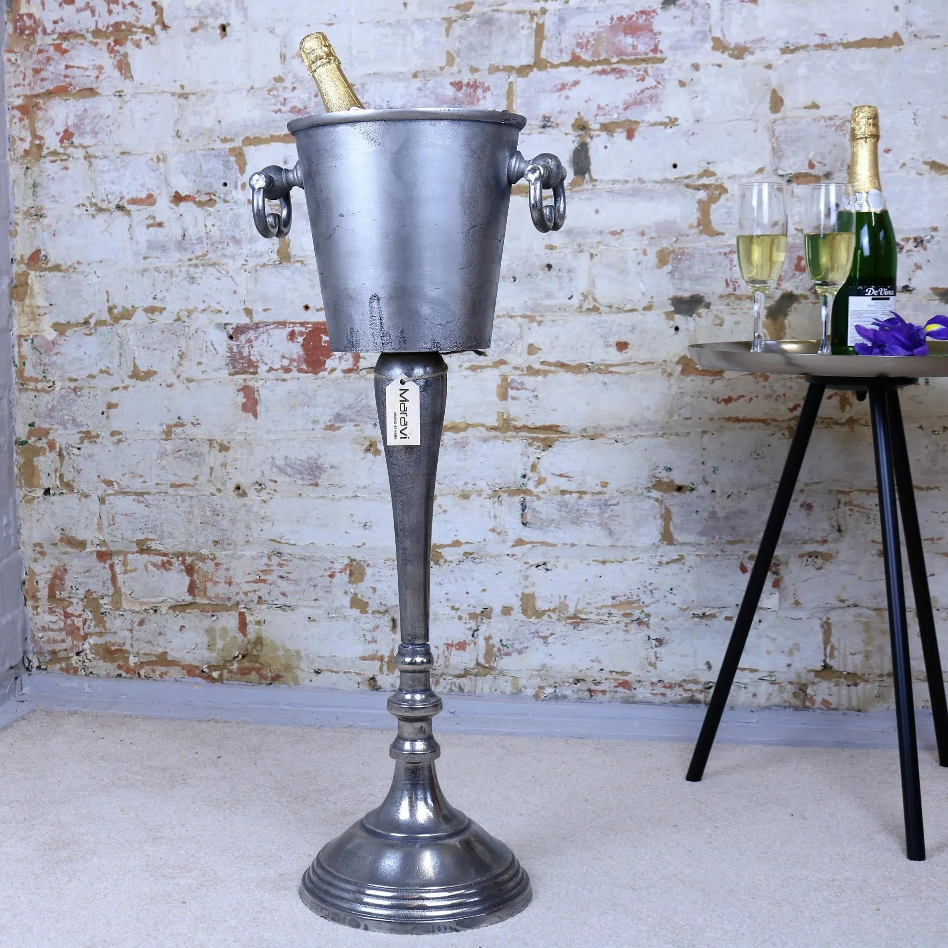 Vintage-style metal ice bucket on a stand with champagne and glasses against a brick wall.