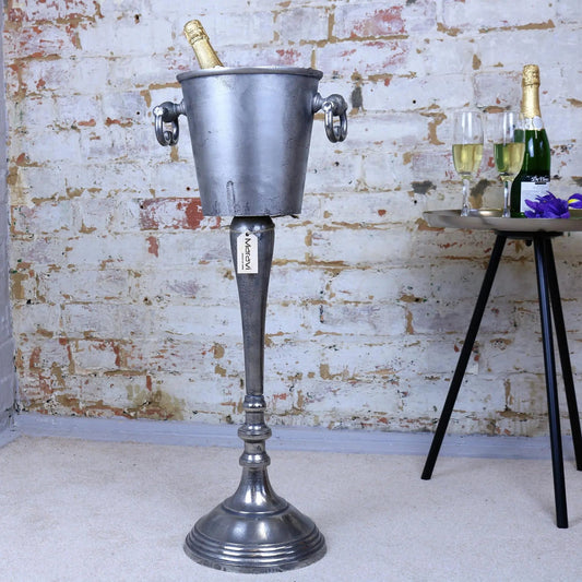 Vintage-style metal ice bucket on a stand with champagne and glasses against a brick wall.