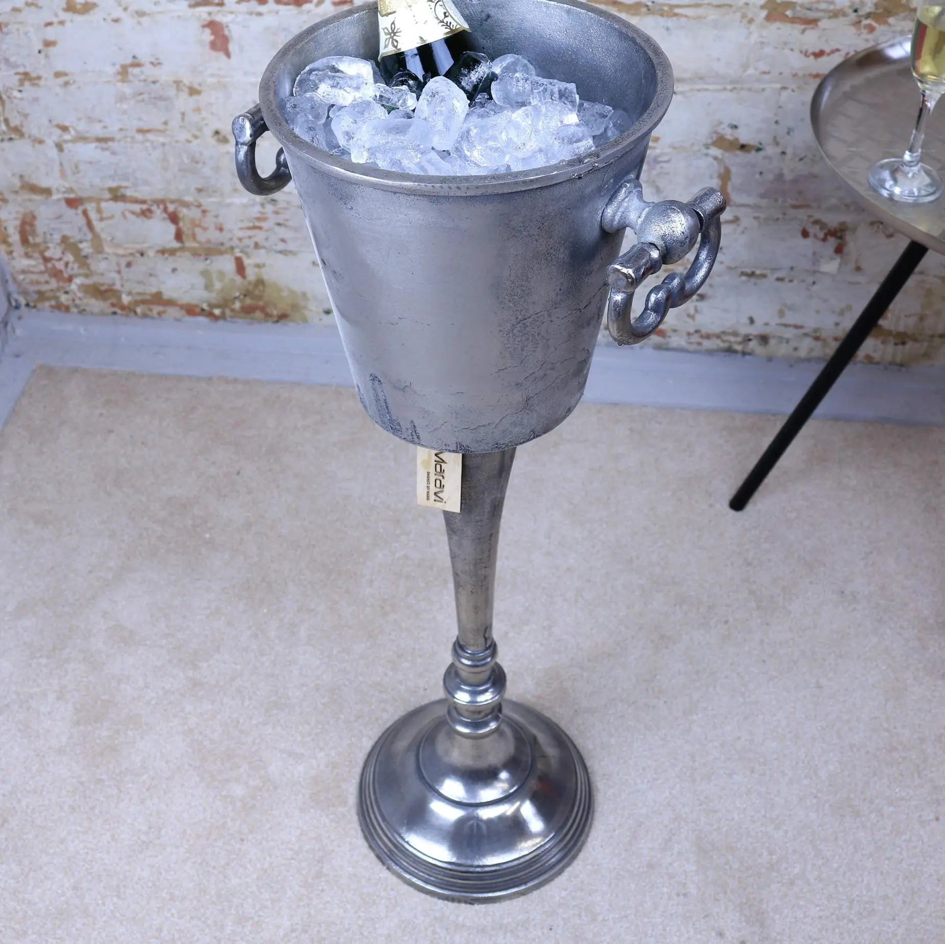 Metallic wine cooler with ice and a bottle of champagne against a rustic brick wall.