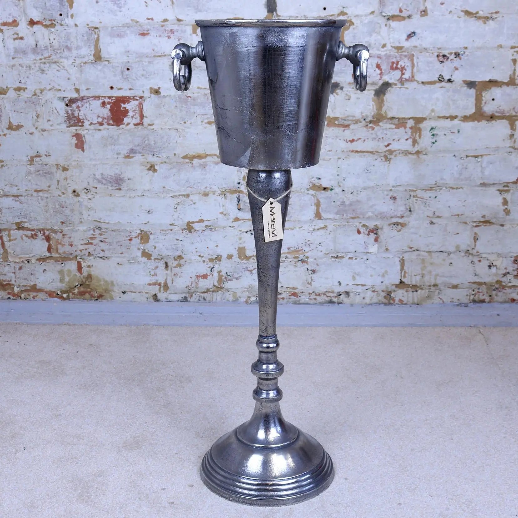 Vintage-style metal ice bucket on a stand against a brick wall.