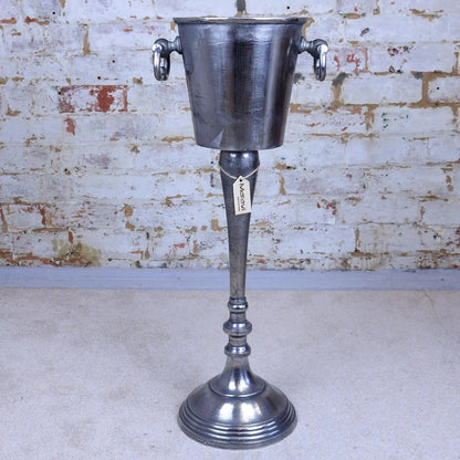 Vintage-style metal ice bucket on a stand against a brick wall.