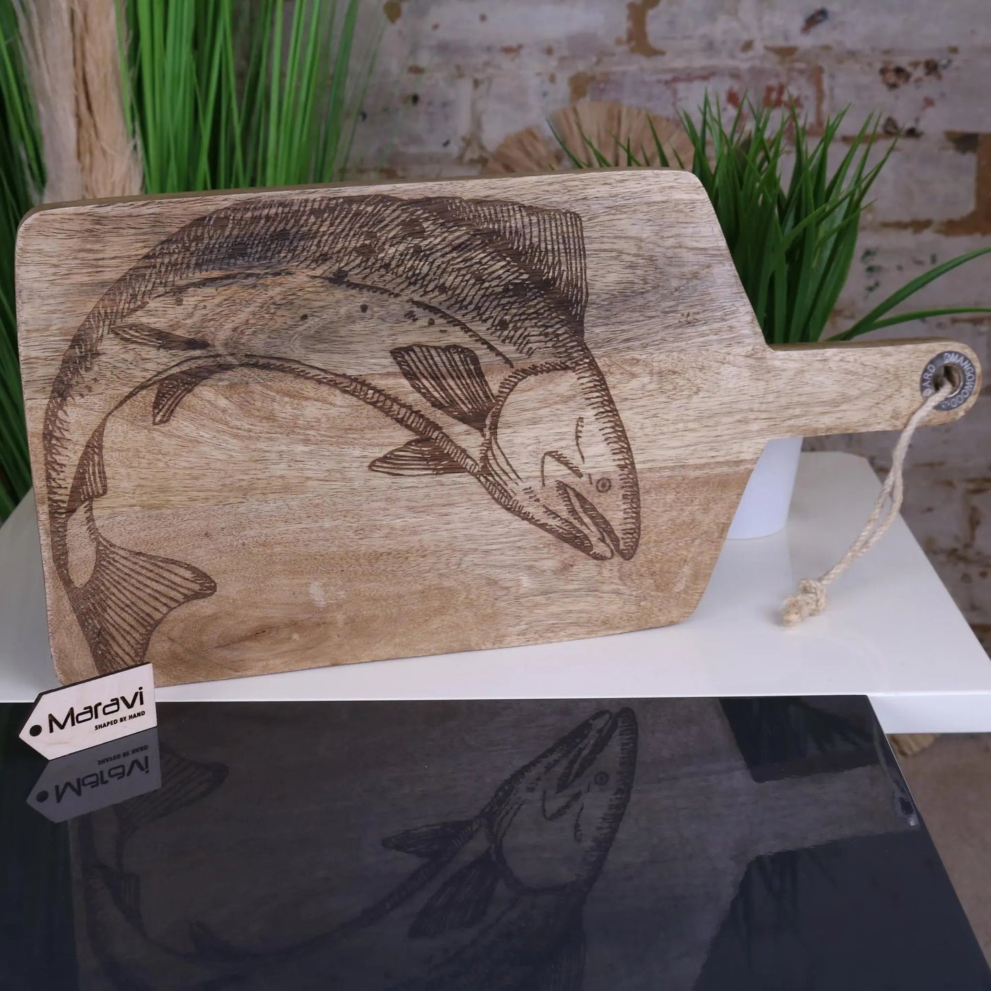 Wooden cutting board with engraved fish design on a white surface with plants in the background