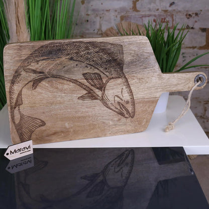 Wooden cutting board with engraved fish design on a white surface with plants in the background