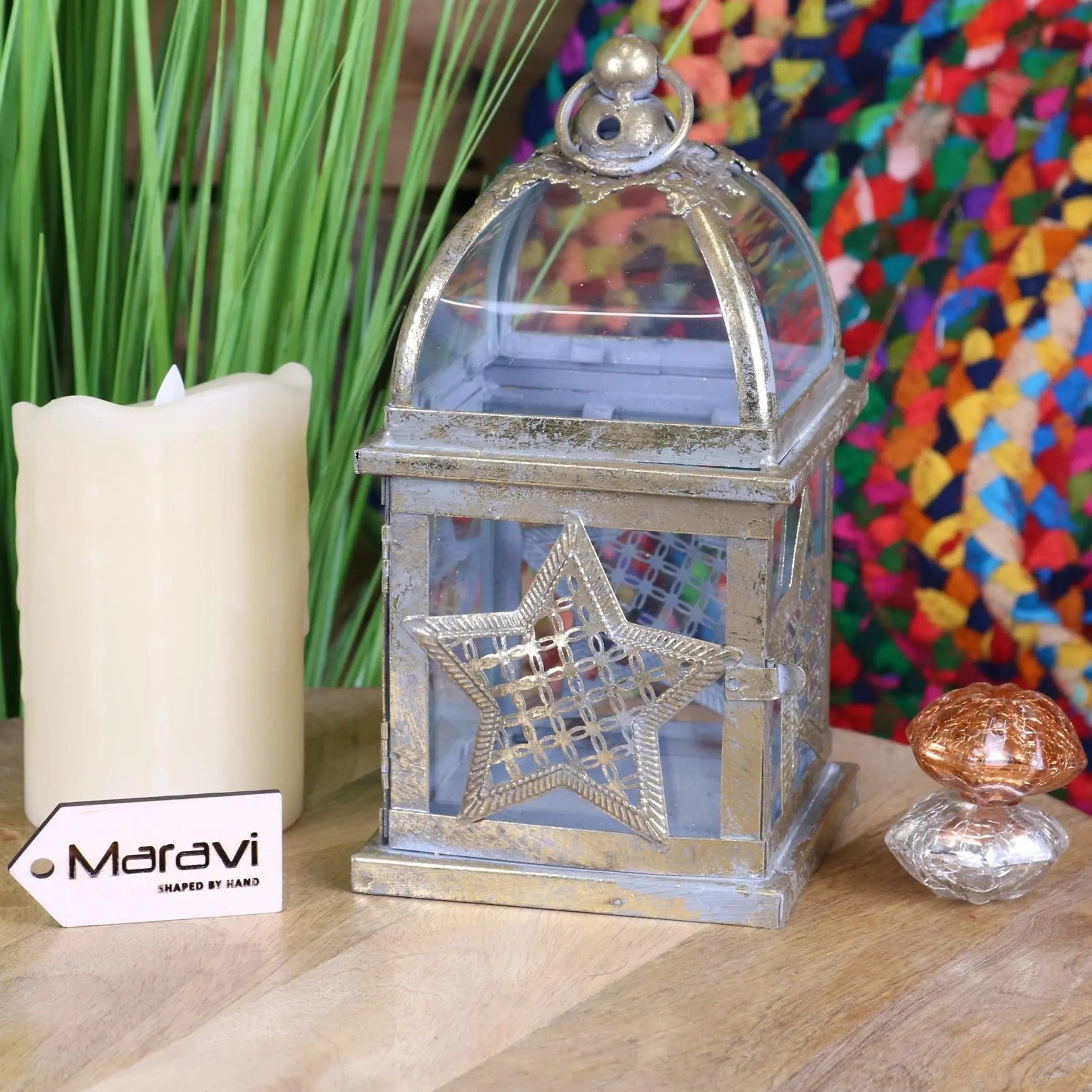 Decorative lantern with star design next to a candle and small object on a wooden surface, with a colorful abstract painting in the background.
