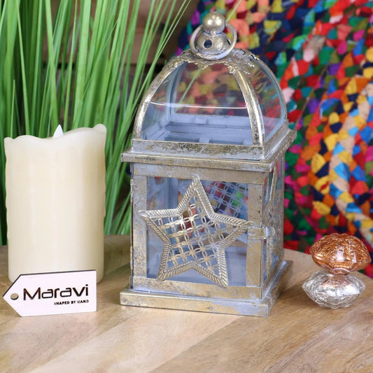 Decorative lantern with star design next to a candle and small object on a wooden surface, with a colorful abstract painting in the background.