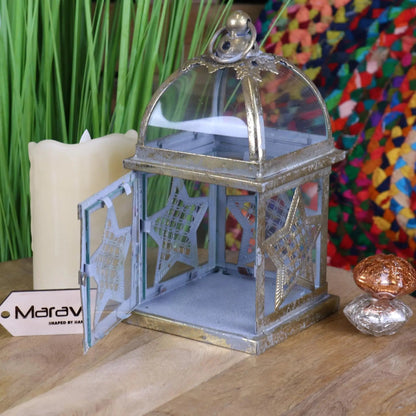 Decorative metal lantern with star designs on a wooden surface, with a colorful fabric and candle in the background.