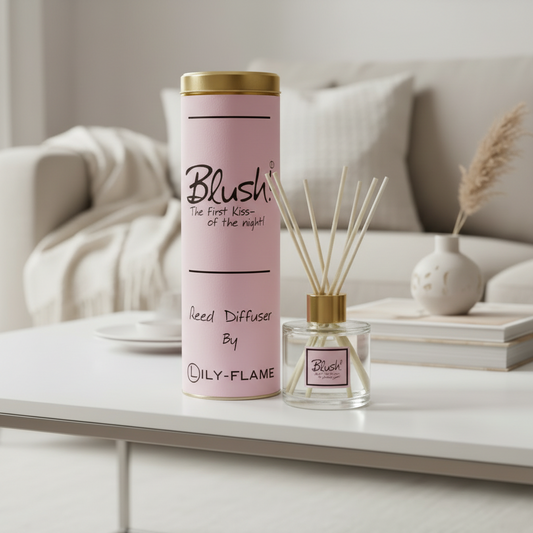 A pink cylindrical reed diffuser canister with a gold lid stands on a white table beside a matching glass reed diffuser with wooden sticks. The packaging and bottle label read 'Blush' and 'By LILY-FLAME'.