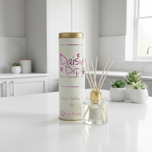 A reed diffuser set on a white countertop in a bright kitchen or bathroom setting. The product includes a tall cylindrical white container with a gold lid and pink 'Daisy Dip' branding, alongside a clear glass reed diffuser bottle with beige reeds and a gold cap.