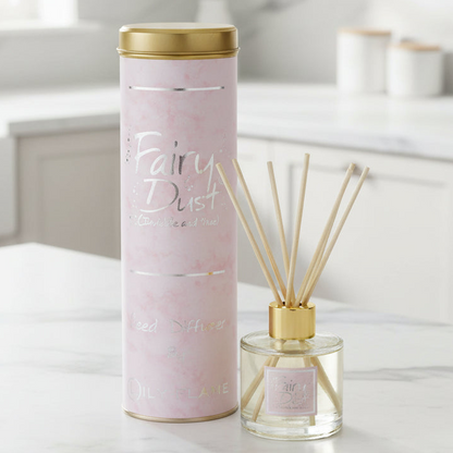 A pink Fairy Dust reed diffuser set by Oily Flame, featuring a tall pink marble-patterned cylindrical tin with a gold lid next to a clear glass bottle with beige reeds and a gold cap, set on a white marble countertop in a bright kitchen.