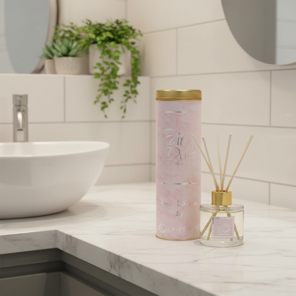 A pink cylindrical diffuser tin with gold trim stands on a marble bathroom counter beside a clear glass reed diffuser bottle with gold cap and wooden reeds. The background shows a white tiled wall, a white vessel sink, and green plants.