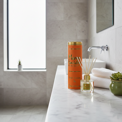 An orange cylindrical diffuser bottle with gold accents stands on a white marble bathroom counter beside a matching reed diffuser, with folded white towels, a small green plant, and a tiled bathroom background.