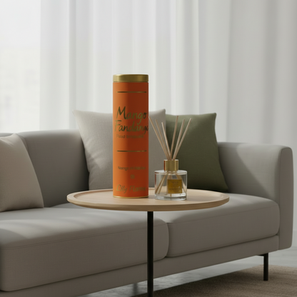 An orange cylindrical reed diffuser bottle with gold lettering stands on a small round wooden side table in a modern living room setting. A matching reed diffuser with glass bottle and wooden reeds sits beside it on the table, with a light gray sofa and neutral pillows in the background.