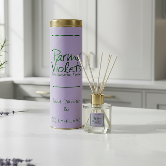 A reed diffuser set on a white countertop in a bright kitchen setting. The product includes a lavender cylindrical tin with a gold lid labeled 'Parmi Violettes' and a clear glass reed diffuser bottle with gold cap and natural reeds, both branded 'ILY-FLAME'.