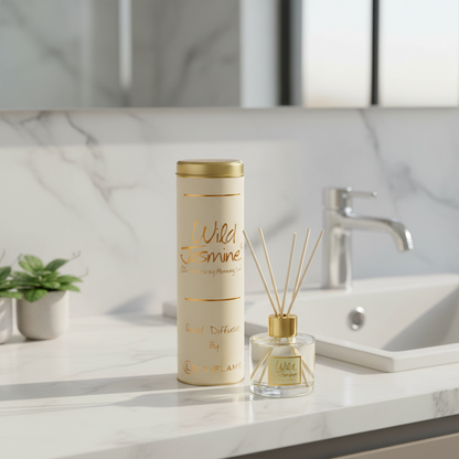 A cylindrical and bottle-style reed diffuser set labeled 'Wild Jasmine' sits on a white marble countertop beside a sink and faucet in a bathroom setting.
