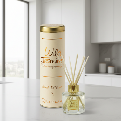 A reed diffuser bottle with natural reeds and a gold cap stands beside its tall cream-colored cylindrical packaging canister on a light marble countertop in a modern kitchen setting.