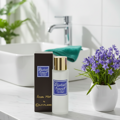 A bottle of Lily Flame Bluebell Forest Room Mist with a gold cap stands next to its dark brown box on a white marble countertop. The background shows a clean, modern bathroom with a white sink, chrome faucet, and a white vase of purple flowers.