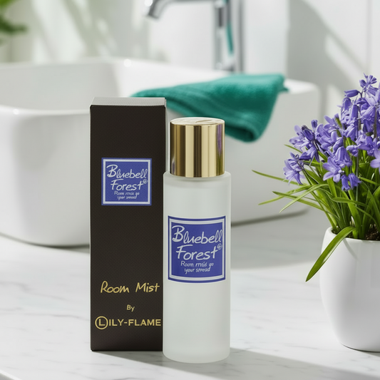 A bottle of Bluebell Forest room mist by LILY-FLAME stands next to its dark brown box on a bathroom counter, with a white sink, a green towel, and a potted purple flower in the background.