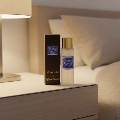 A bottle of Bluebell Forest Room Mist by Oily-Flame stands on a bedside table next to its black box, with a softly lit lamp and bed in the background.
