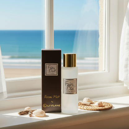 A bottle of Fairy Dust Room Mist by Lyl-Flame stands next to its dark brown box on a sunlit windowsill with a beach and ocean view in the background. The bottle is frosted with a gold cap and a silver label, and the box is dark brown with gold lettering.