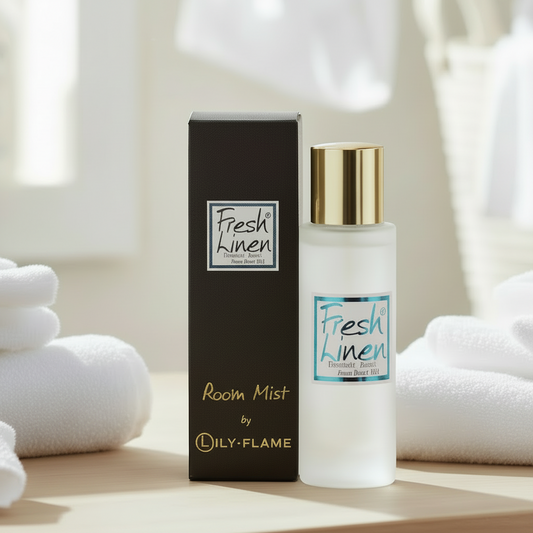 A bottle of Fresh Linen room mist by LILY-FLAME stands next to its black packaging box on a light wooden surface, styled with folded white towels in a bright, soft-focus bathroom setting.