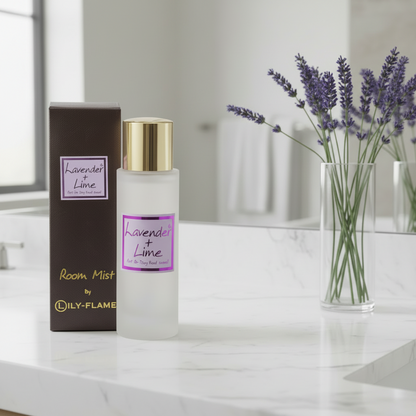 A frosted glass bottle of Lily-Flame Lavender + Lime Room Mist with a gold cap stands next to its dark brown box on a white marble countertop. A glass vase with lavender sprigs is in the background.