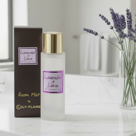 A frosted glass room mist bottle with a gold cap stands next to its dark brown box labeled 'Lavender + Lime' by LILY FLAME. The scene is styled on a white marble surface with a glass vase of lavender in the background.
