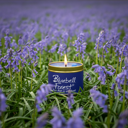 A lit blue candle tin labeled 'Bluebell Forest' sits in a field of purple bluebells, with the flowers surrounding the product in a natural outdoor setting.