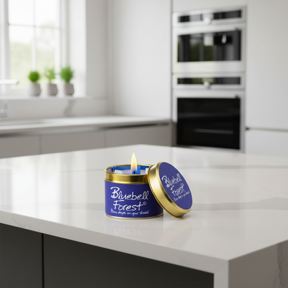A lit blue candle in a round tin with a gold lid resting beside it on a white kitchen countertop. The label reads 'Bluebell Forest' and the lid has matching branding.