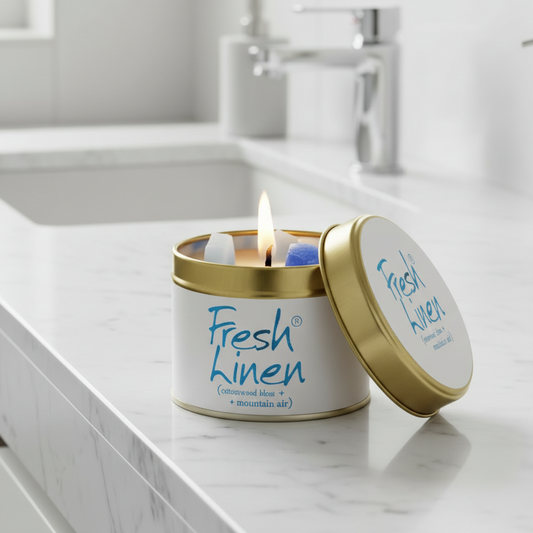 A lit candle in a white tin with a gold lid, labeled 'Fresh Linen' and placed on a white marble bathroom counter with a blurred sink in the background.