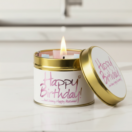 A lit candle in a round gold metal tin with a white label reading 'Happy Birthday!' in pink script, shown on a light marble-like surface with the lid leaning beside it.